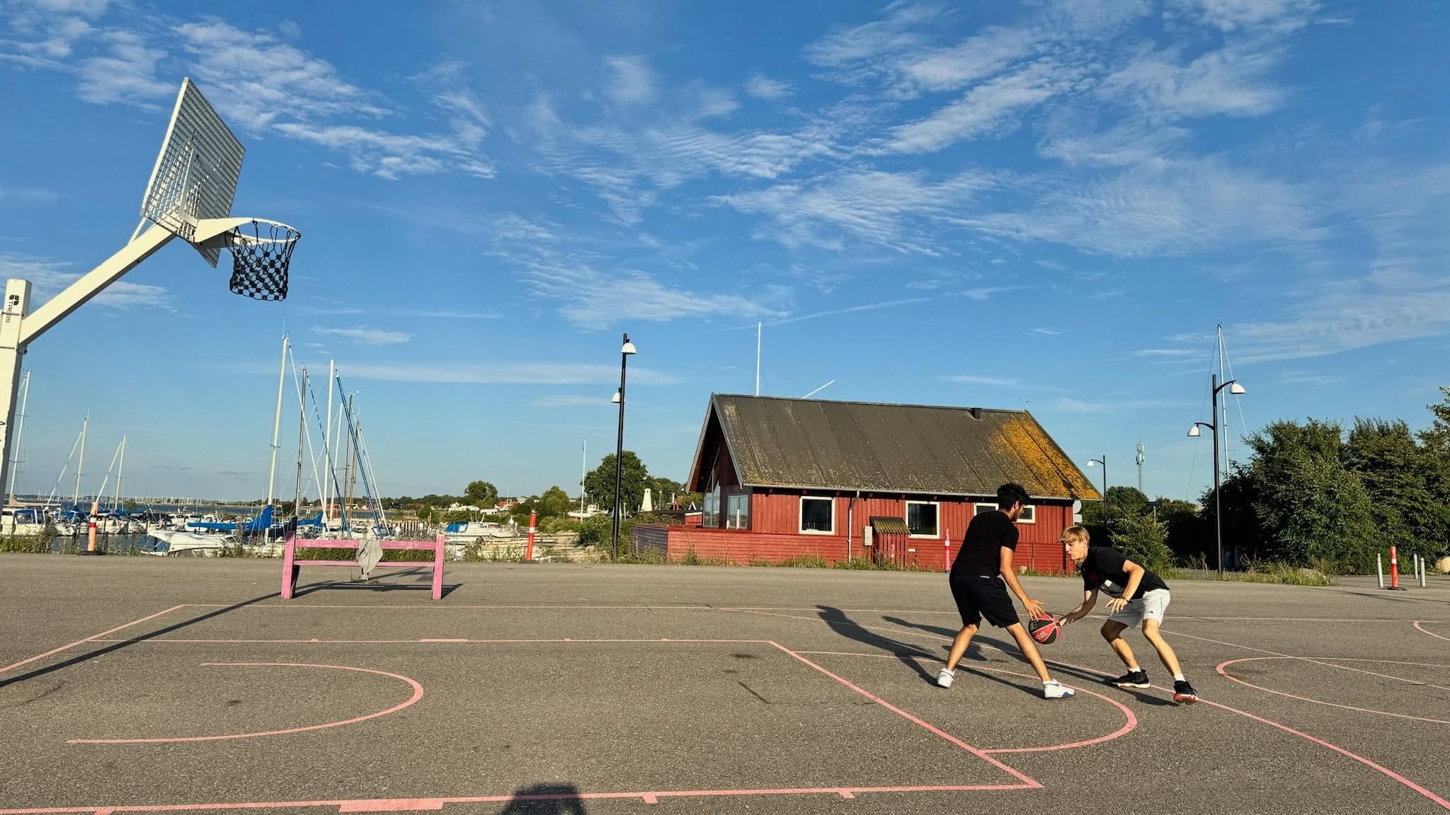 To unge spiller basketball på en udendørs bane ved havnen en sommerdag